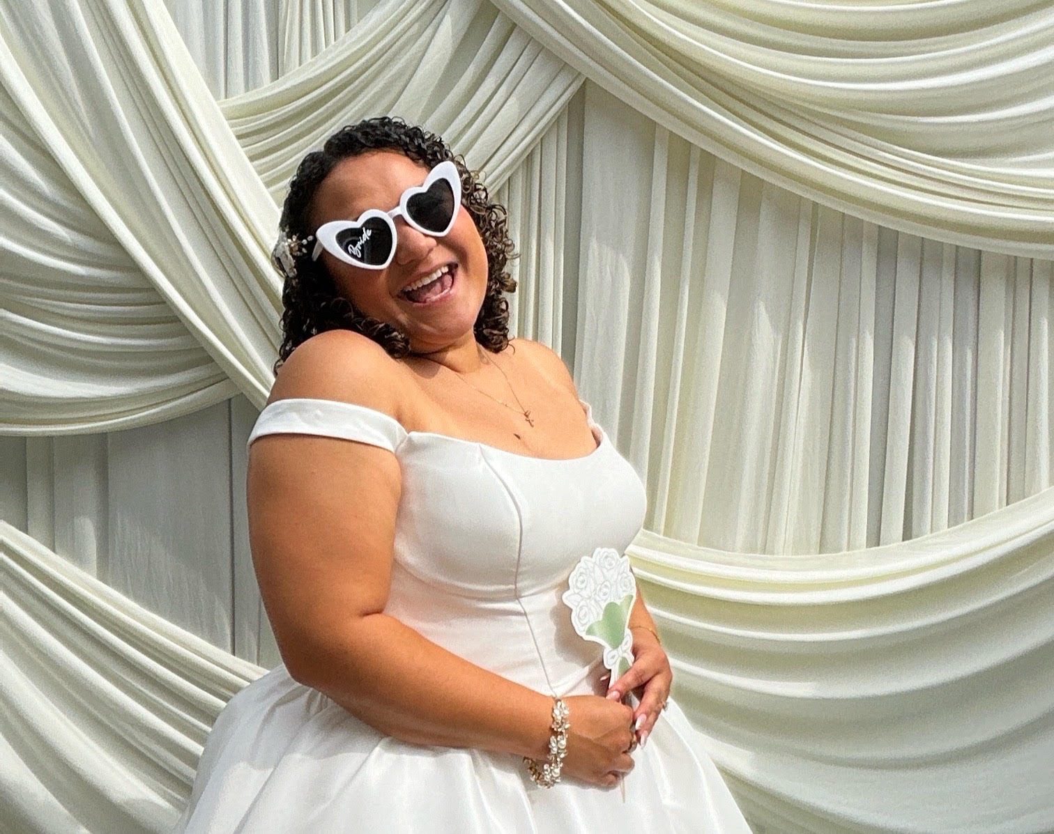 A bride in a white gown wearing heart shaped sunglasses in front of ivory draping
