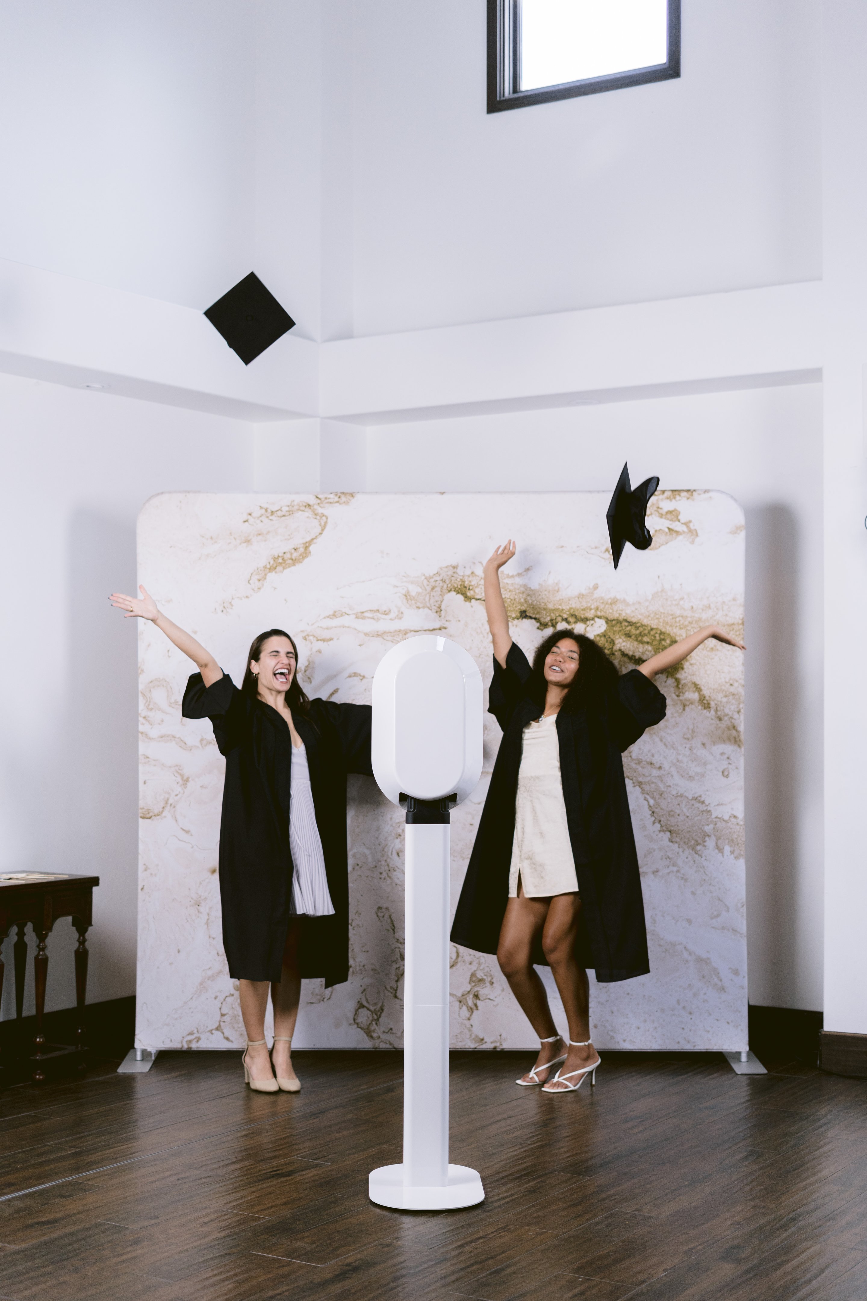 Two graduates in black gowns toss their caps beside a white photo booth and marble print backdrop