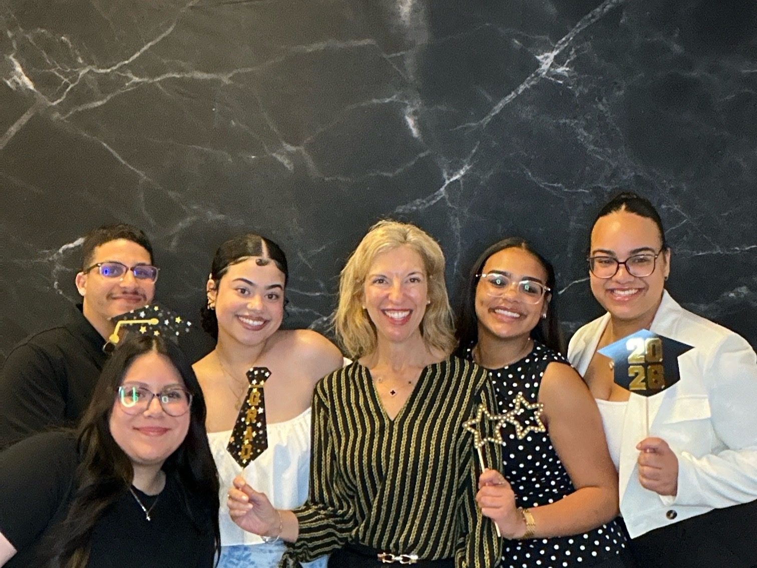 Six guests posing with graduation props, including a Grad tie and a 2026 cap sign, before a black marble backdrop