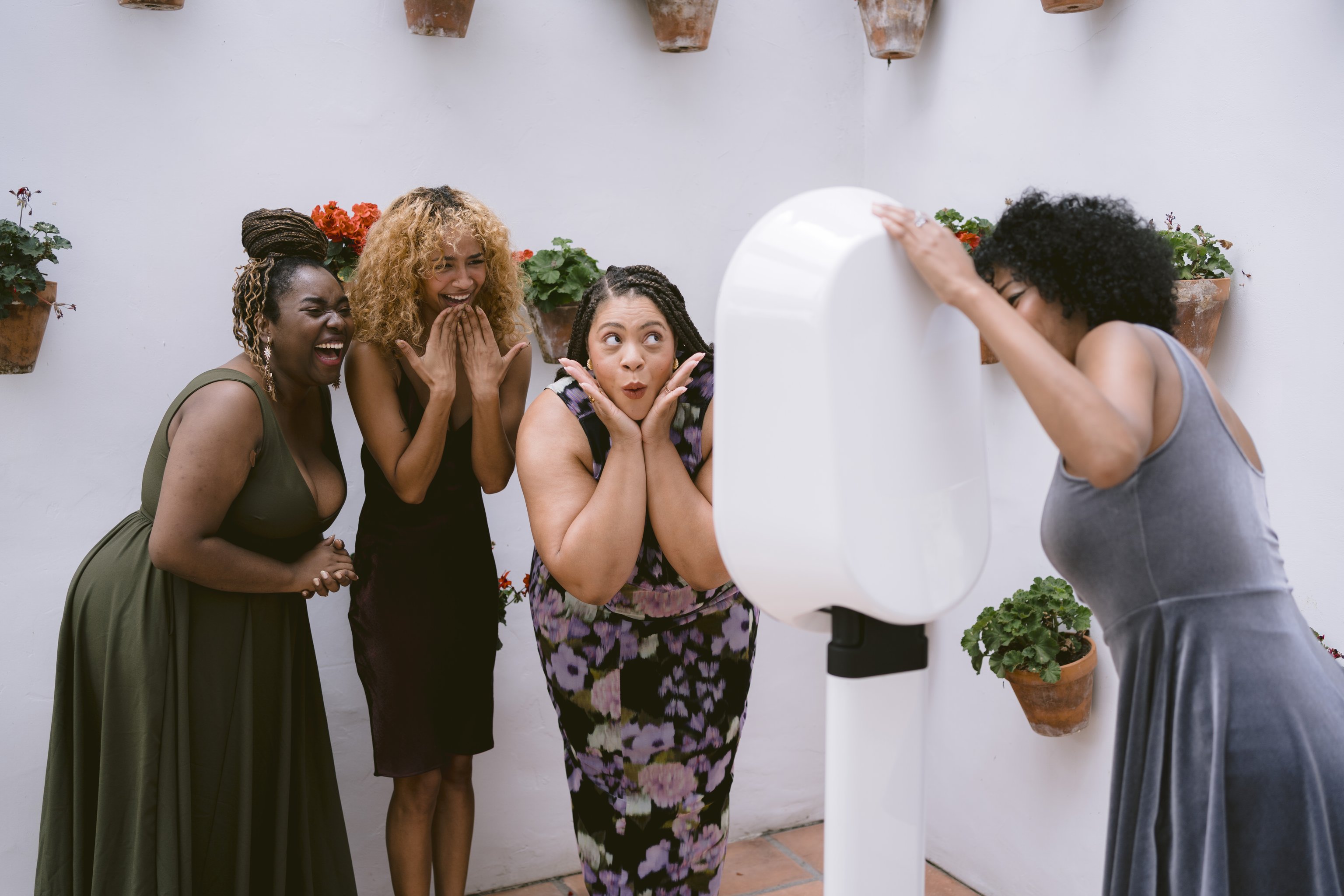 A woman leans in to a white photo booth as three friends react in a plant-lined courtyard