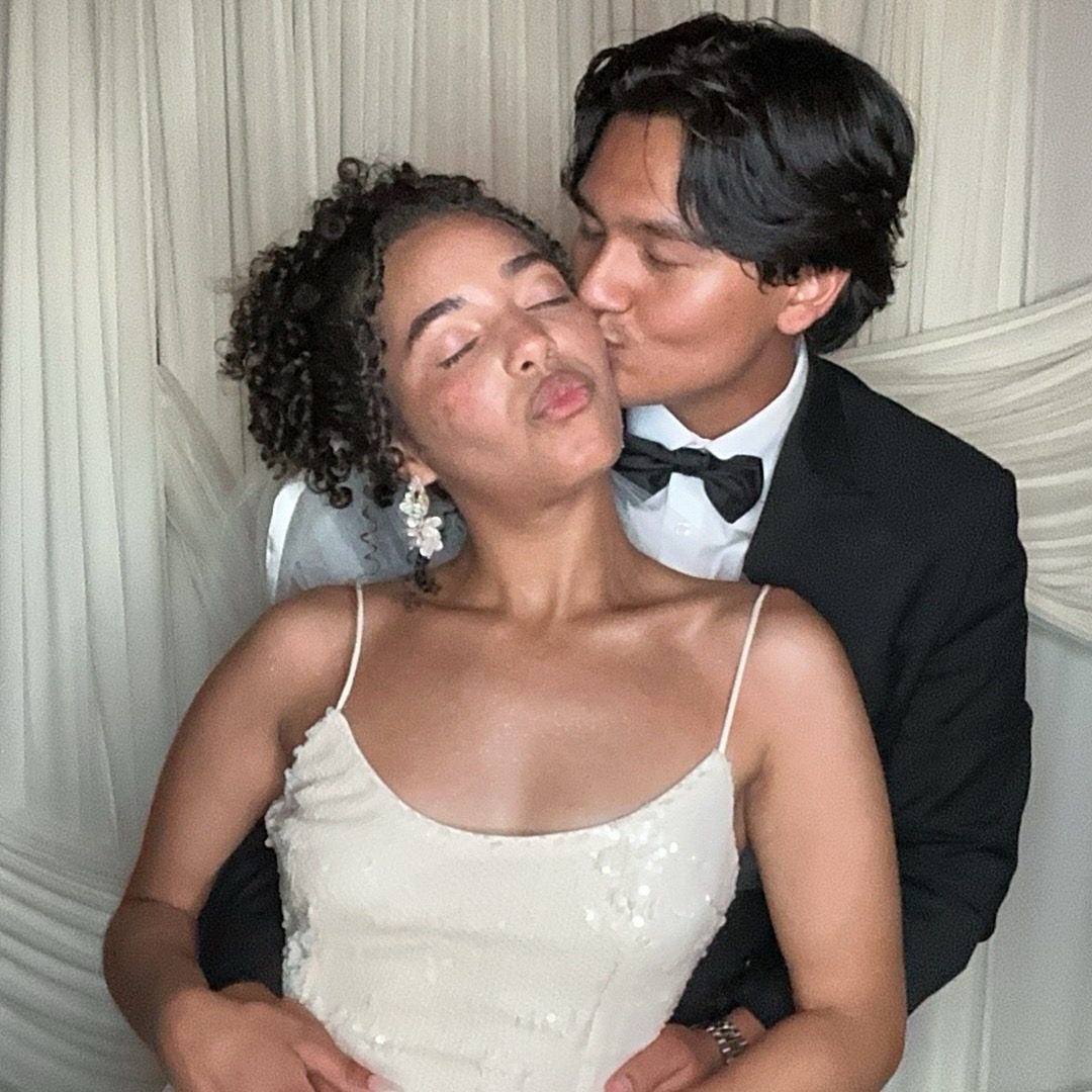 A couple in front of an ivory draped backdrop, one kissing the other on the cheek