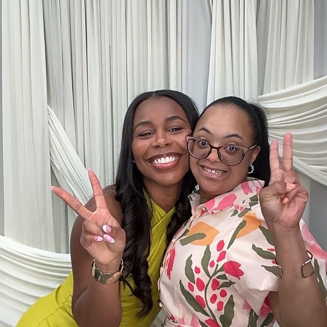 Two women flash peace signs in front of a white draped backdrop, one in a floral print shirt