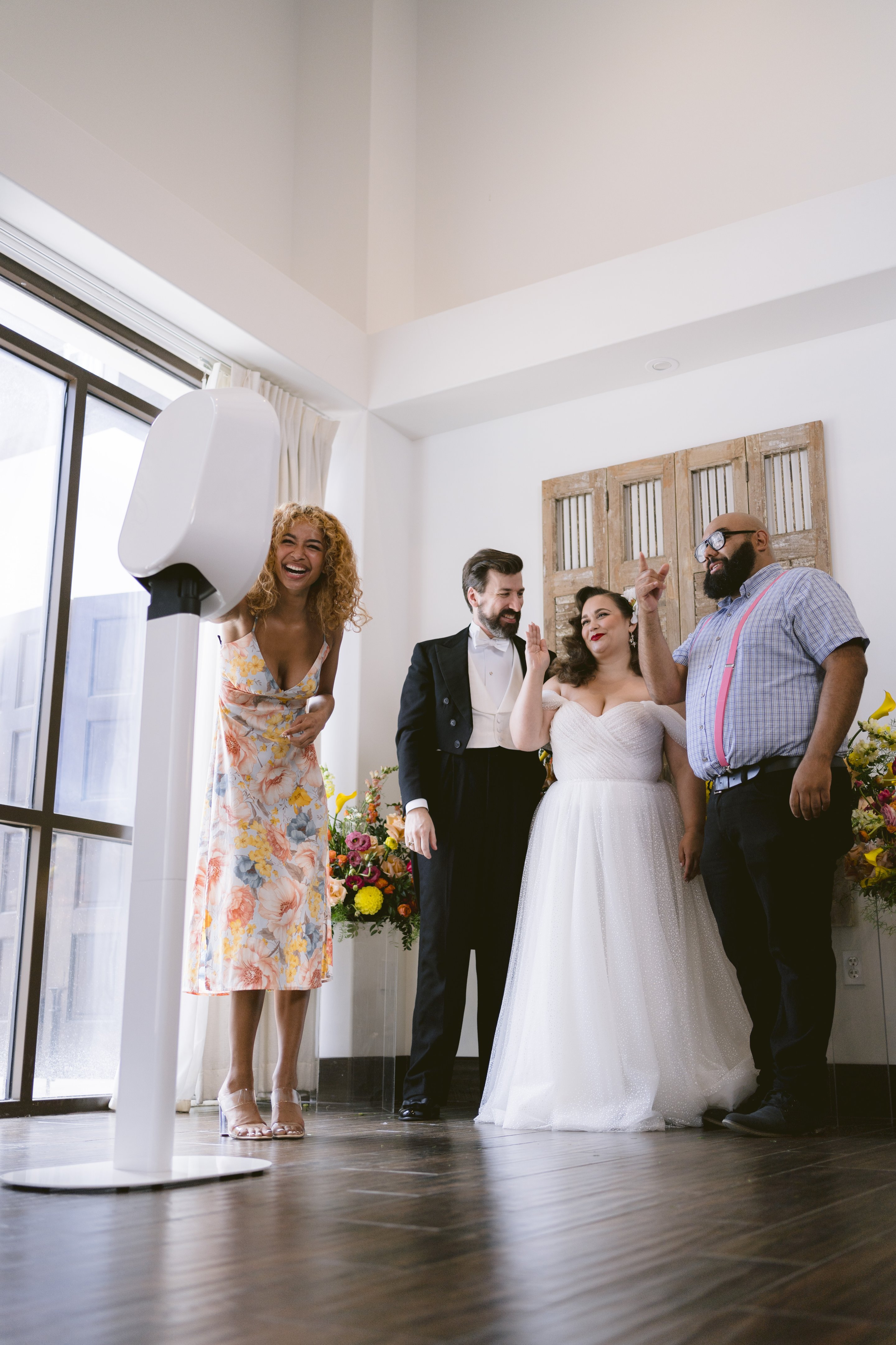 A woman laughs at a white photo booth by tall windows as a bride, groom and guest pose nearby